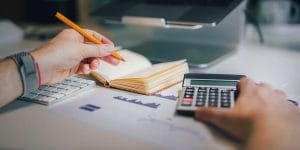 A photo of a person sitting at a desk with a pencil in one hand and the other pushing buttons on a calculator.