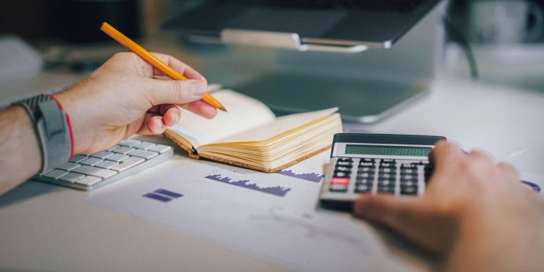 A photo of a person sitting at a desk with a pencil in one hand and the other pushing buttons on a calculator.