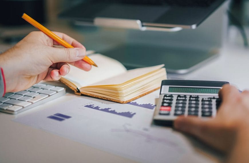 A photo of a person sitting at a desk with a pencil in one hand and the other pushing buttons on a calculator.